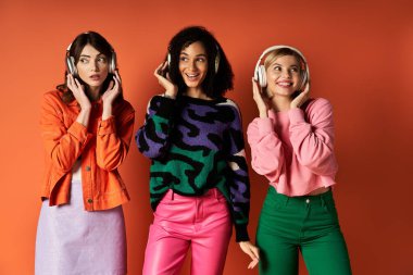 A trio of stylish, multicultural women stand united on an orange background, symbolizing friendship and cultural diversity. clipart