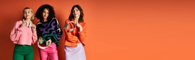 Three young, beautiful women with headphones striking a pose in front of a bold orange wall. clipart