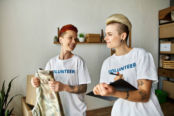 Молодая пара лесбиянок в волонтерских футболках стоят вместе в комнате, занимаясь благотворительностью.