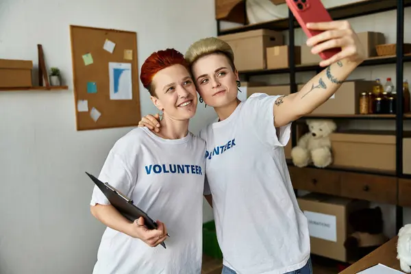 stock image Young lesbian couple in volunteer shirts capturing a moment of connection with a selfie in a room.