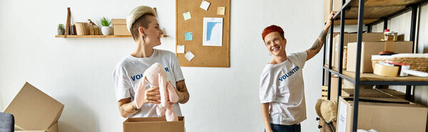 A young lesbian couple in volunteer shirts, standing in a room filled with boxes, working together for charity.