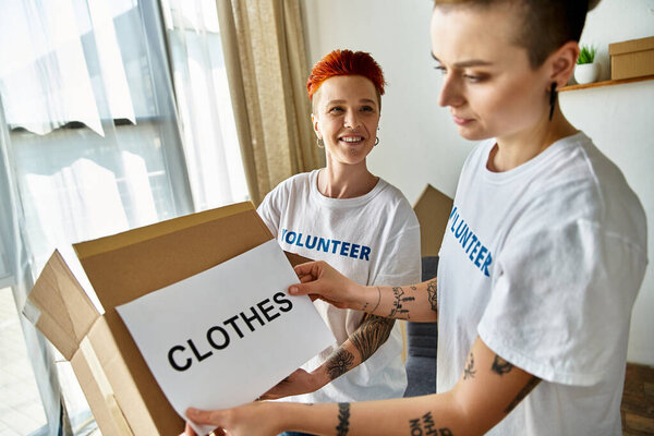 Молодая пара лесбиянок в футболках волонтеров с коробкой пожертвованной одежды.
