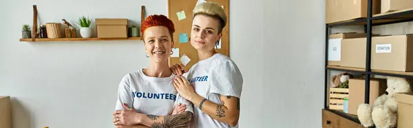 stock image Young lesbian couple in volunteer t-shirts working on a charity project side by side in a room.