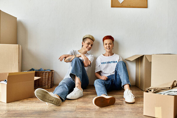 A young lesbian couple in volunteer shirts sit on the floor amid numerous boxes, engaged in charity work together.