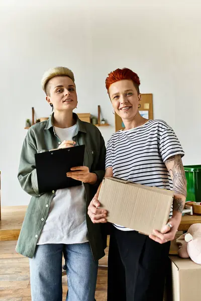 stock image Young lesbian couple stand side by side, holding boxes filled with donations for charity work.