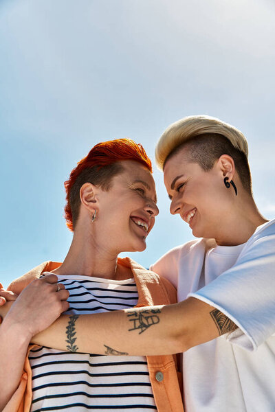 A young lesbian couple proudly standing side by side holding a rainbow flag outdoors.