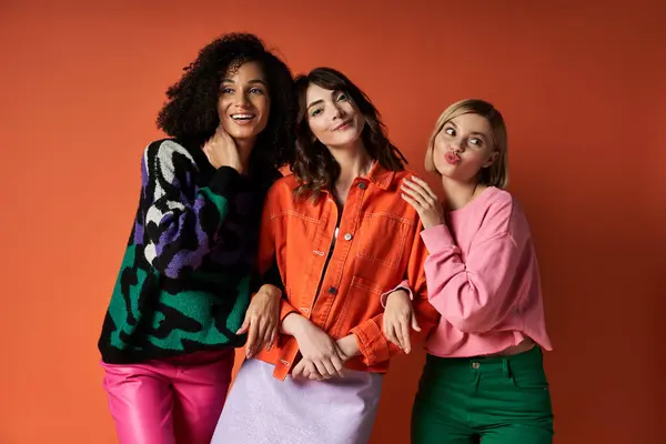 stock image A trio of young, multicultural women in vibrant clothes pose together against an orange background.