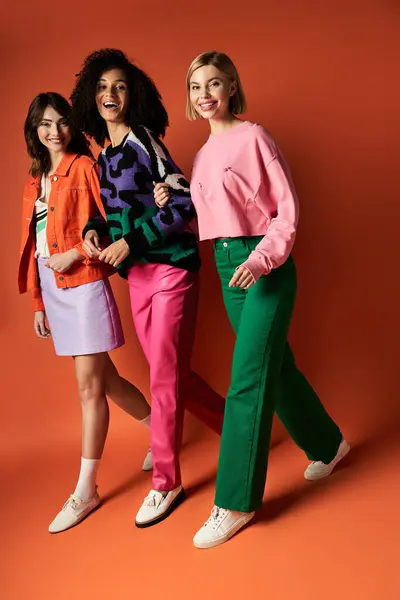 stock image Three young, multicultural women stand in stylish vibrant clothes against an orange backdrop, exuding friendship and diversity.