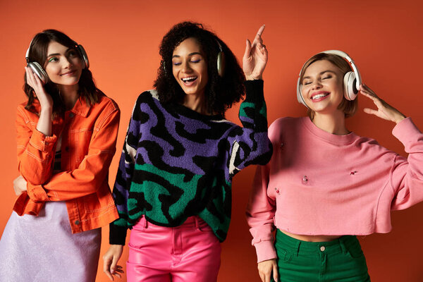 Three young beautiful multicultural women in stylish outfits posing together on an orange background.
