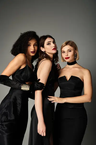 stock image Three young women, each in elegant black dresses, standing gracefully together on a grey background.