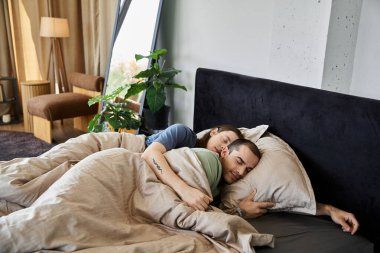 Genç eşcinsel bir çift, modern bir yatak odası ortamında birbirlerine sokulup samimiyet ve sevgi gösteriyorlar..