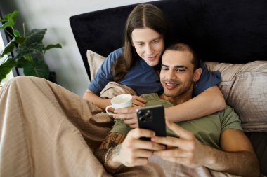 A young gay couple cuddles in bed, one man holding a coffee mug while the other looks at his phone. clipart