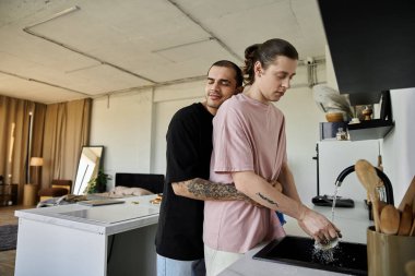 A young gay couple enjoys a moment of intimacy while washing dishes in their modern home. clipart