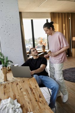 A young gay couple spends quality time together in their modern home, one man working on a laptop while the other brings him a drink. clipart