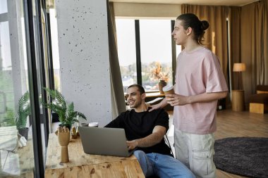 Two young men relax in their modern home. One works on a laptop as the other brings him coffee. clipart