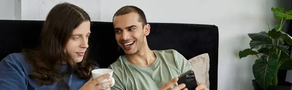 stock image A young gay couple relaxes in bed, sharing a quiet moment together. One partner holds a cup, while the other looks at a smartphone.