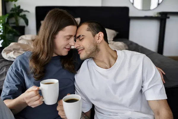 stock image A young gay couple enjoys a quiet morning together in their bedroom, sharing a cup of coffee and a tender moment.