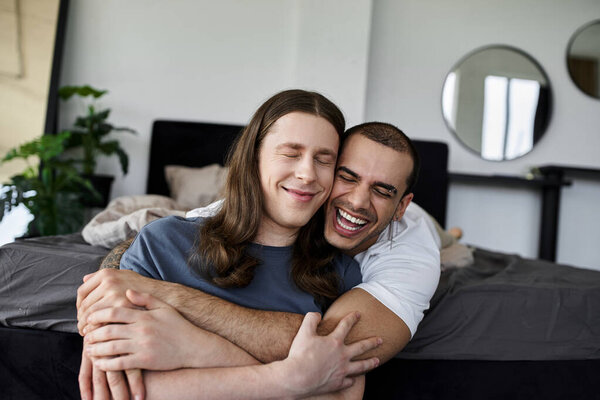 A young gay couple smiles and laughs as they embrace each other on a bed in a bedroom.