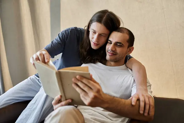 stock image Two young men cuddle on a couch, one reading a book while the other rests their head on his shoulder.