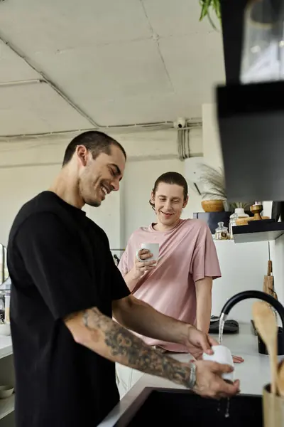 stock image Two young men, in a modern home, enjoy a casual morning moment as one washes dishes while the other holds a mug.