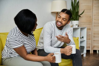 An African American couple chats on yellow couch, enjoying tea. clipart