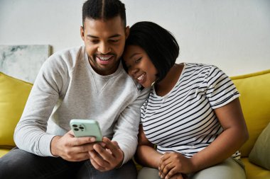Sevgi dolu bir Afrikalı Amerikalı çift kanepede akıllı telefona bakarken birlikte gülüyor..
