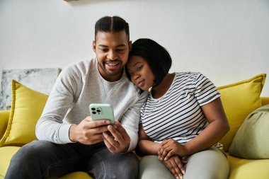 A loving African American couple relaxes on a yellow couch together, looking at a phone. clipart