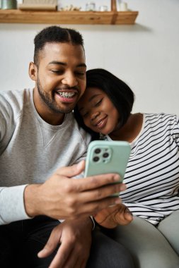 Afro-Amerikalı bir çift koltukta sarılıyor, birlikte telefona bakarken gülüyorlar..