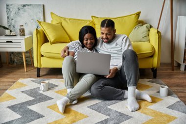 Sevgi dolu bir Afrikalı Amerikalı çift oturma odasındaki halıya dizüstü bilgisayar kullanarak rahatlıyor..