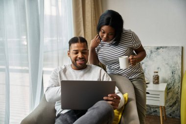Afro-Amerikalı çift evde rahat, dizüstü bilgisayar kullanıyor ve içkinin tadını çıkarıyor..