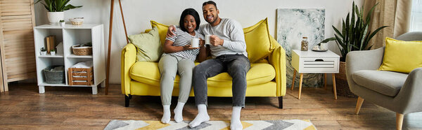 Афро-американская пара отдыхает дома на жёлтом диване, наслаждаясь горячим напитком.