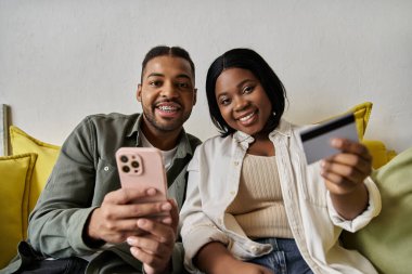 Sevgi dolu bir Afro-Amerikan çifti evde online alışveriş yapmak için telefon ve kredi kartı kullanıyor..