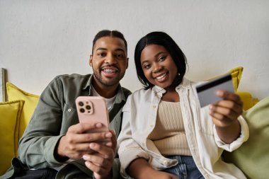 A loving African American couple uses a phone and credit card to shop online at home. clipart