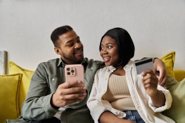 Bir Afrikalı Amerikalı çift online alışveriş yaparken kredi kartı ve akıllı telefonuyla gülümsüyor..