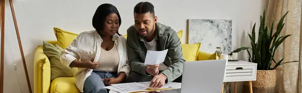 stock image An African American couple sits on a yellow sofa, reviewing paperwork and using a laptop.