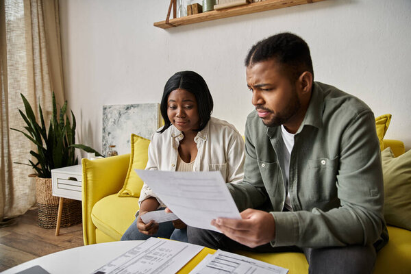 A loving African American couple sits on a yellow couch, reviewing documents together at home.