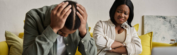 Обеспокоенная афроамериканская пара сидит на диване, мужчина расстроен, женщина смотрит на него.