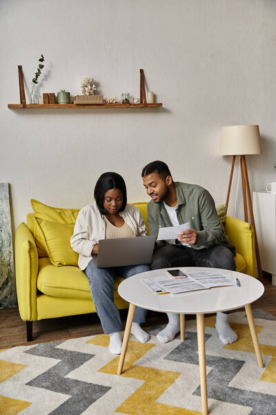A couple lounges on yellow couch, sharing laptop.