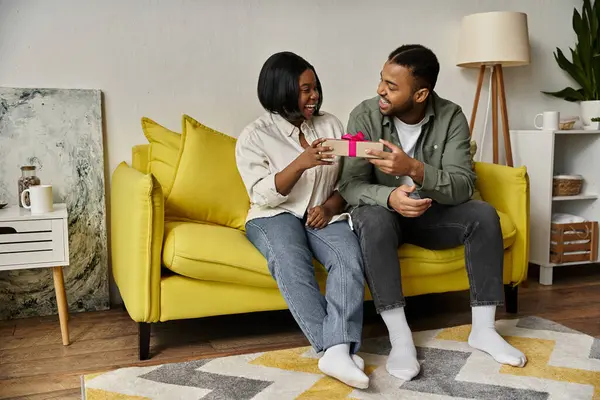 stock image A happy African American couple sits on a yellow sofa at home, exchanging a gift.