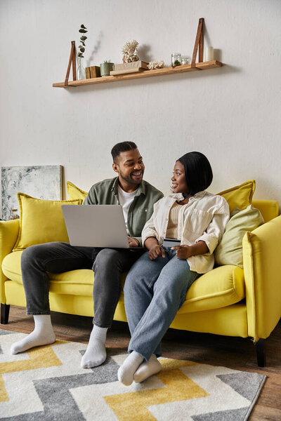 A loving African American couple smiles and chats while relaxing on a yellow sofa in their home.
