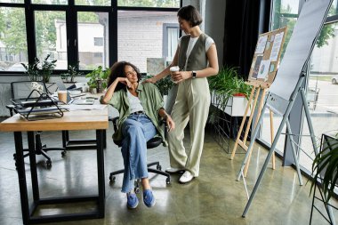 İki kadın bir ofis ortamında, biri oturuyor, biri ayakta, bir projede birlikte çalışıyorlar..