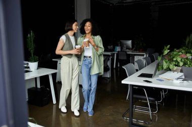 İki kadın, lezbiyen bir çift, kahve fincanlarını tutarken ofis ortamında birlikte gülüyorlar..