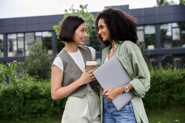 İki kadın iş kıyafeti giymiş, gülümsüyor, ellerinde dizüstü bilgisayar ve kahve fincanı var..