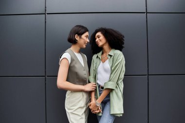 Birbirine aşık iki kadın birbirine yakın durup gri bir duvara gülümsüyor..