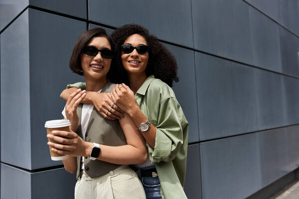 Две женщины в солнечных очках обнимаются перед современным зданием.
