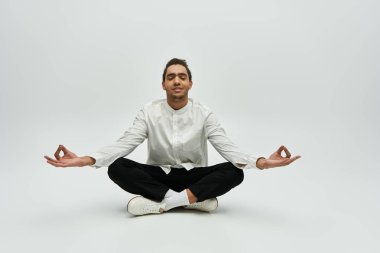 Beyaz gömlekli ve siyah pantolonlu bir Afro-Amerikan erkek model, gözleri kapalı meditasyon yapıyor, gri arka planda bacak bacak üstüne atarak oturuyor..