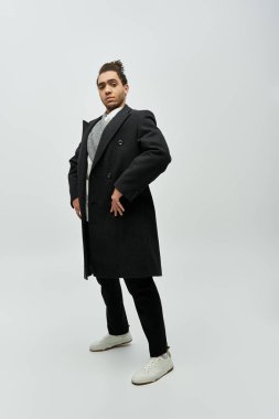 Tarz sahibi bir Afrikalı Amerikalı siyah bir ceket ve beyaz bir spor ayakkabıyla gri bir arka planda duruyor..