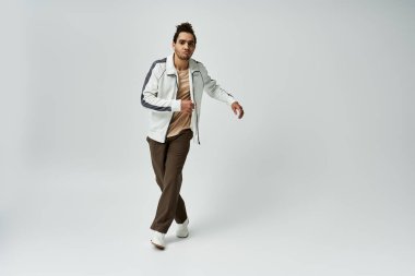 Genç bir Afrikalı Amerikalı, gri arka planda şık beyaz bir ceket ve kahverengi bir pantolon giyerek güvenle poz veriyor..