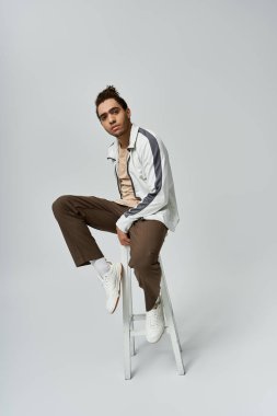 Beyaz ceketli ve kahverengi pantolonlu Afrikalı genç bir adam gri bir fona karşı taburede poz veriyor..