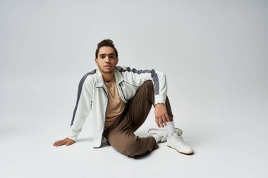 Beyaz ceketli ve kahverengi pantolonlu genç bir Afro-Amerikalı gri arka planda poz veriyor..